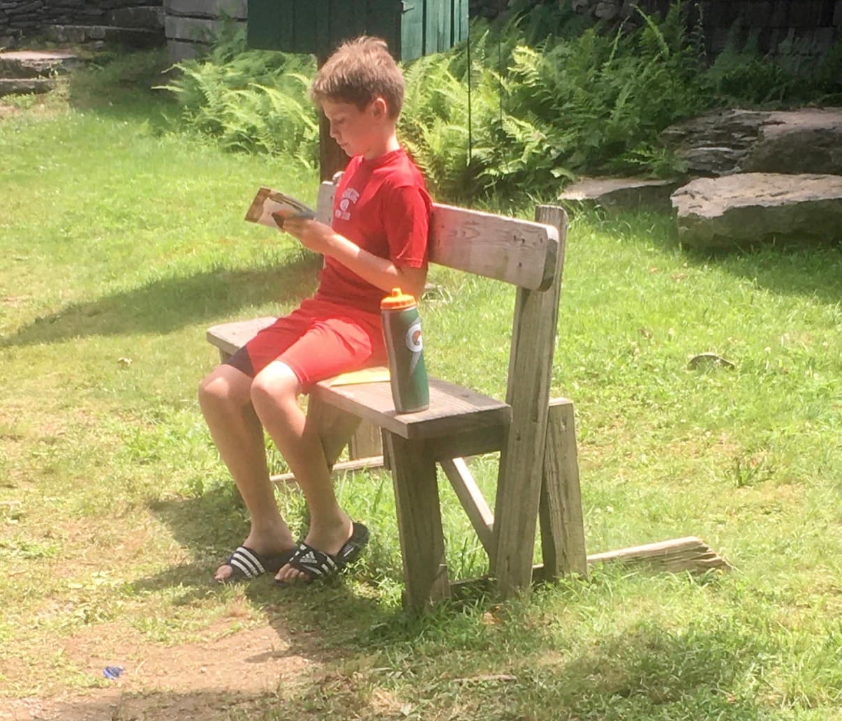 A young boy reading a book on a wooden bench at camp